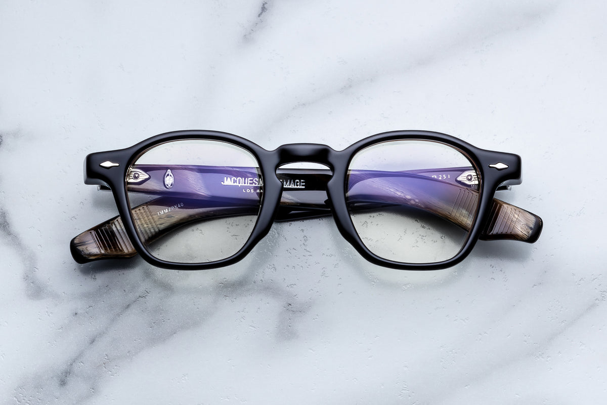 Black-rimmed Jacques Marie Mage Zephirin spectacles with sculpted, patterned brown and gray temples lying on a white marble surface.