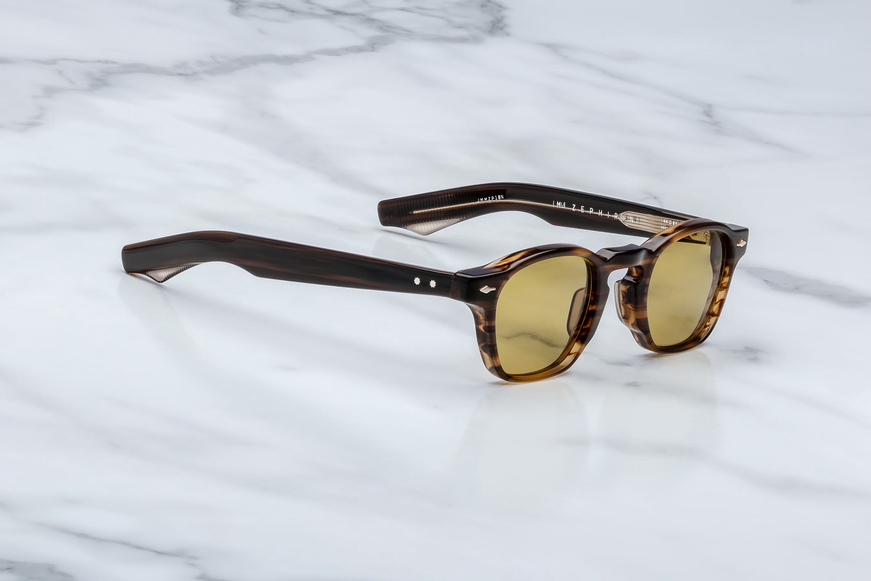 Jacques Marie Mage sunglasses in dark tortoise acetate with yellow tinted lenses and translucent striped temple tips, shown at an angle on a light marble surface.