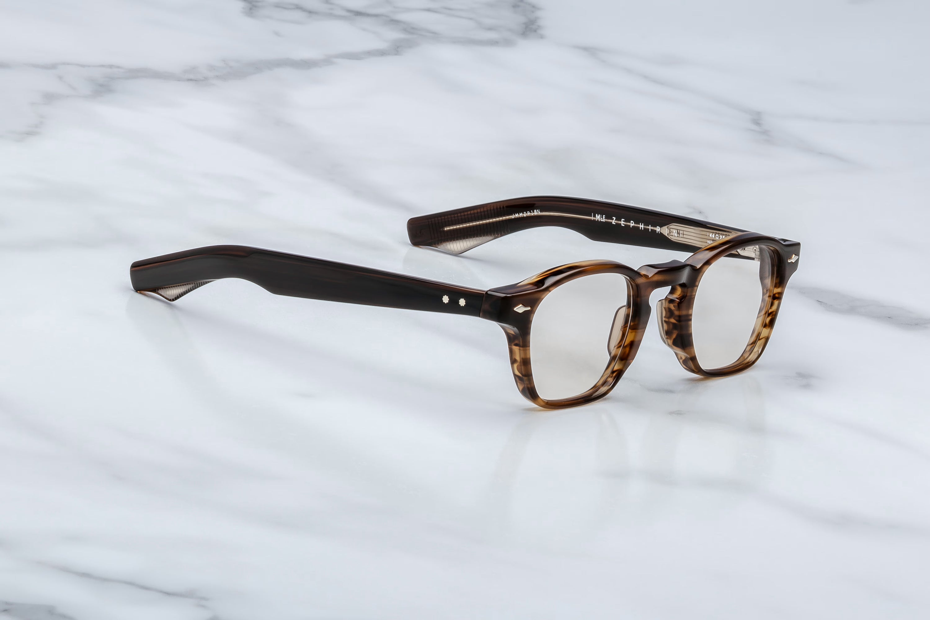 Jacques Marie Mage optical glasses in dark tortoise acetate with clear lenses and translucent striped temple tips, shown at an angle on a light marble surface.