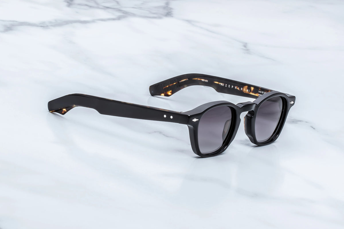 Black-framed Jacques Marie Mage Zephirin spectacles with a tortoise shell inner temple design, shown at an angle on a marble surface.