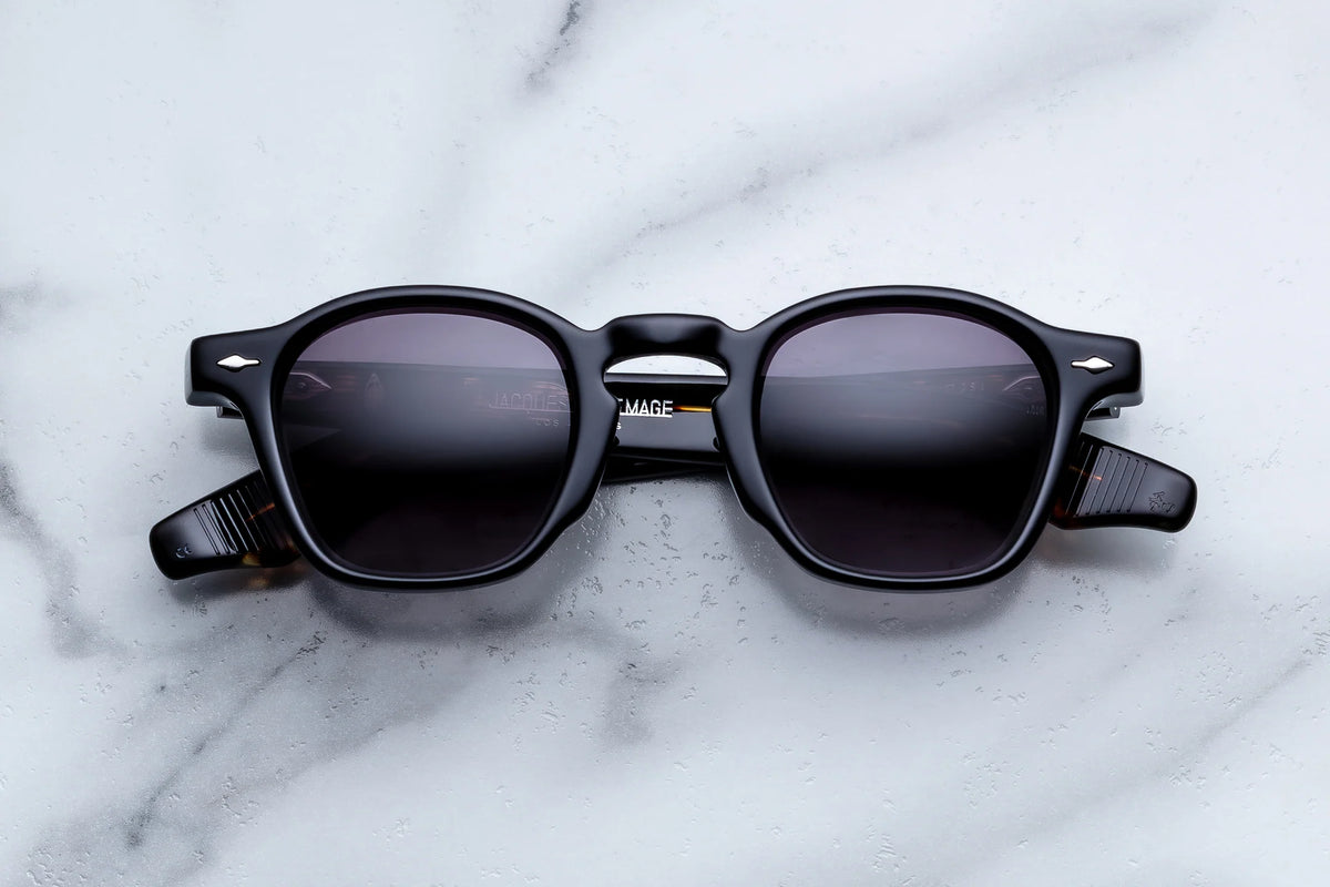Black-framed Jacques Marie Mage Zephirin spectacles with dark lenses laid flat on a white marble surface, featuring hand-finished sculpted temple construction.