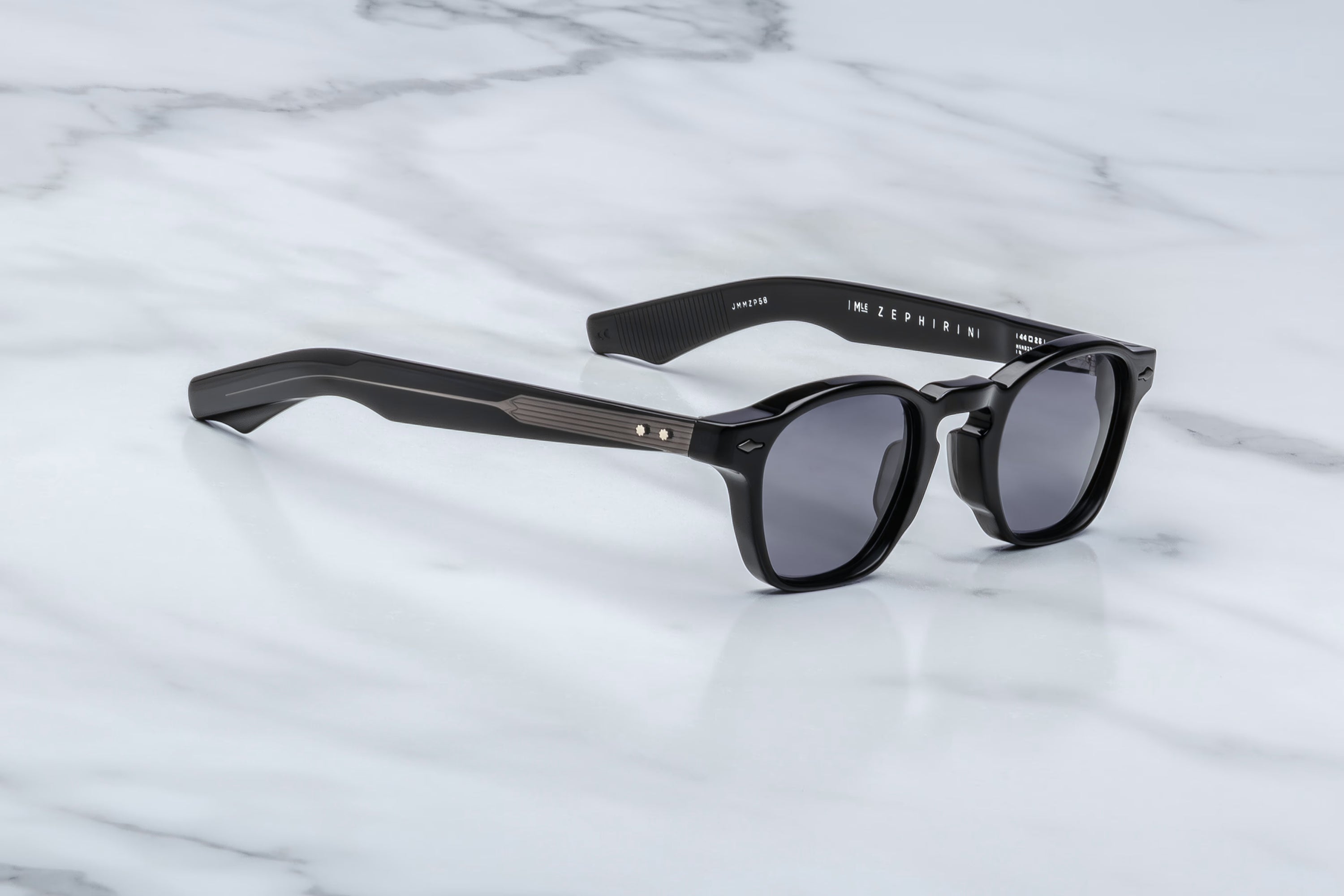 Black acetate sunglasses with grey-tinted lenses, silver arrowhead rivets, and visible wire-core detailing along the temples, shown at an angled side view on a light marble surface.
