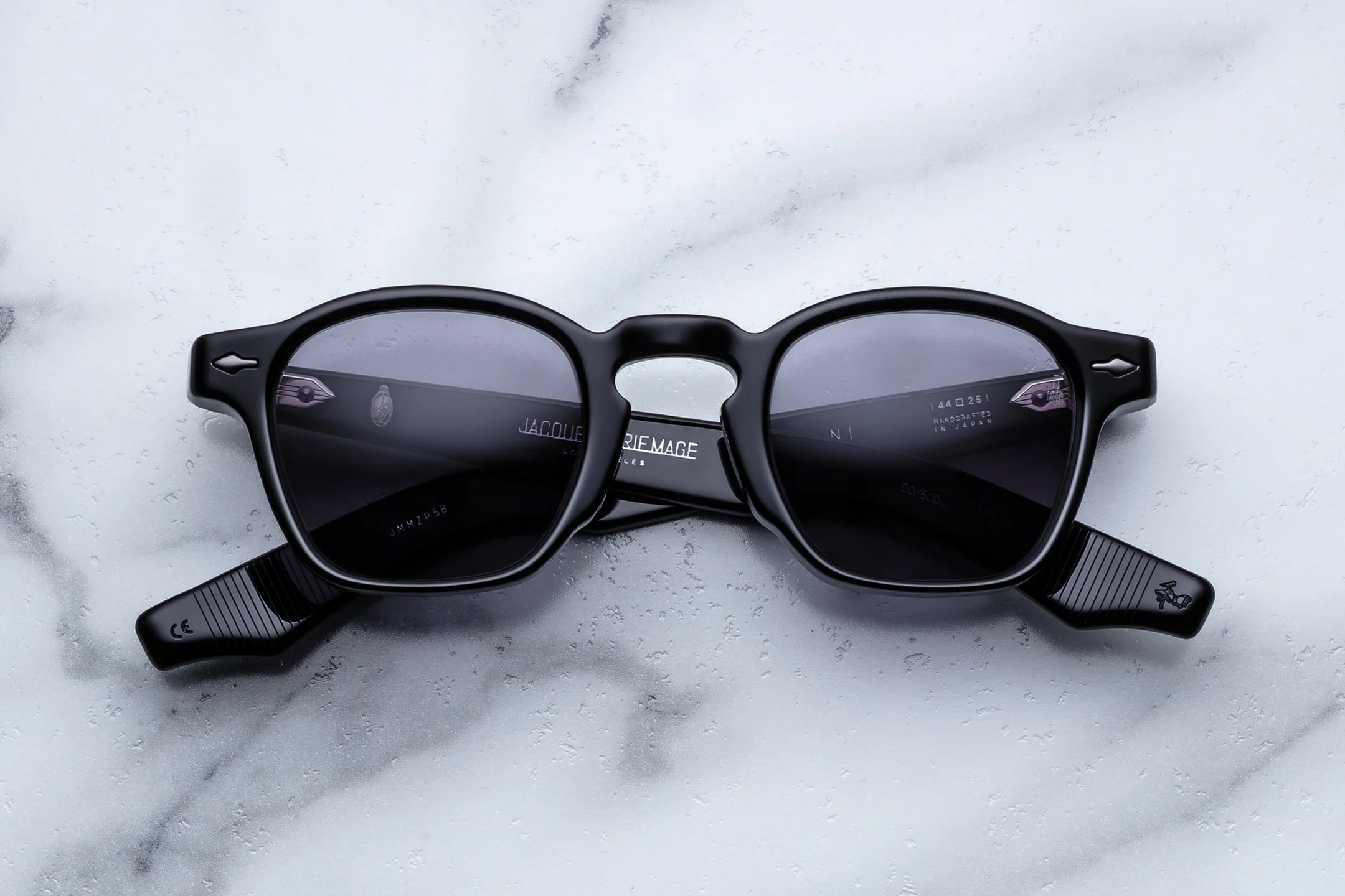 Black acetate sunglasses with grey-tinted lenses, silver arrowhead rivets, and exposed wire-core temple detailing, shown front-facing on a light marble surface.
