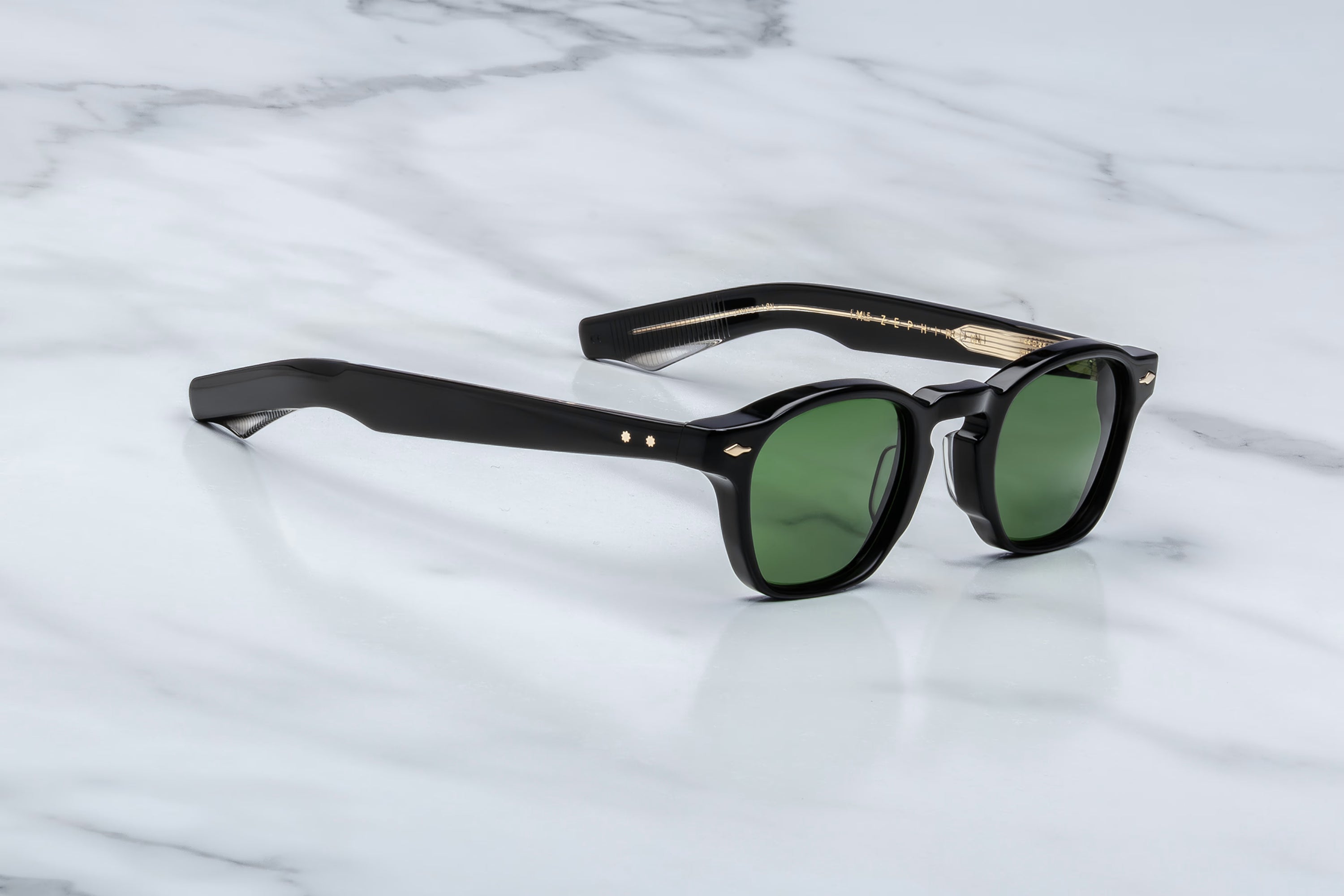 Black acetate sunglasses with green lenses, gold rivet accents, and visible wire-core temple detailing, shown at an angled side view on a light marble surface.