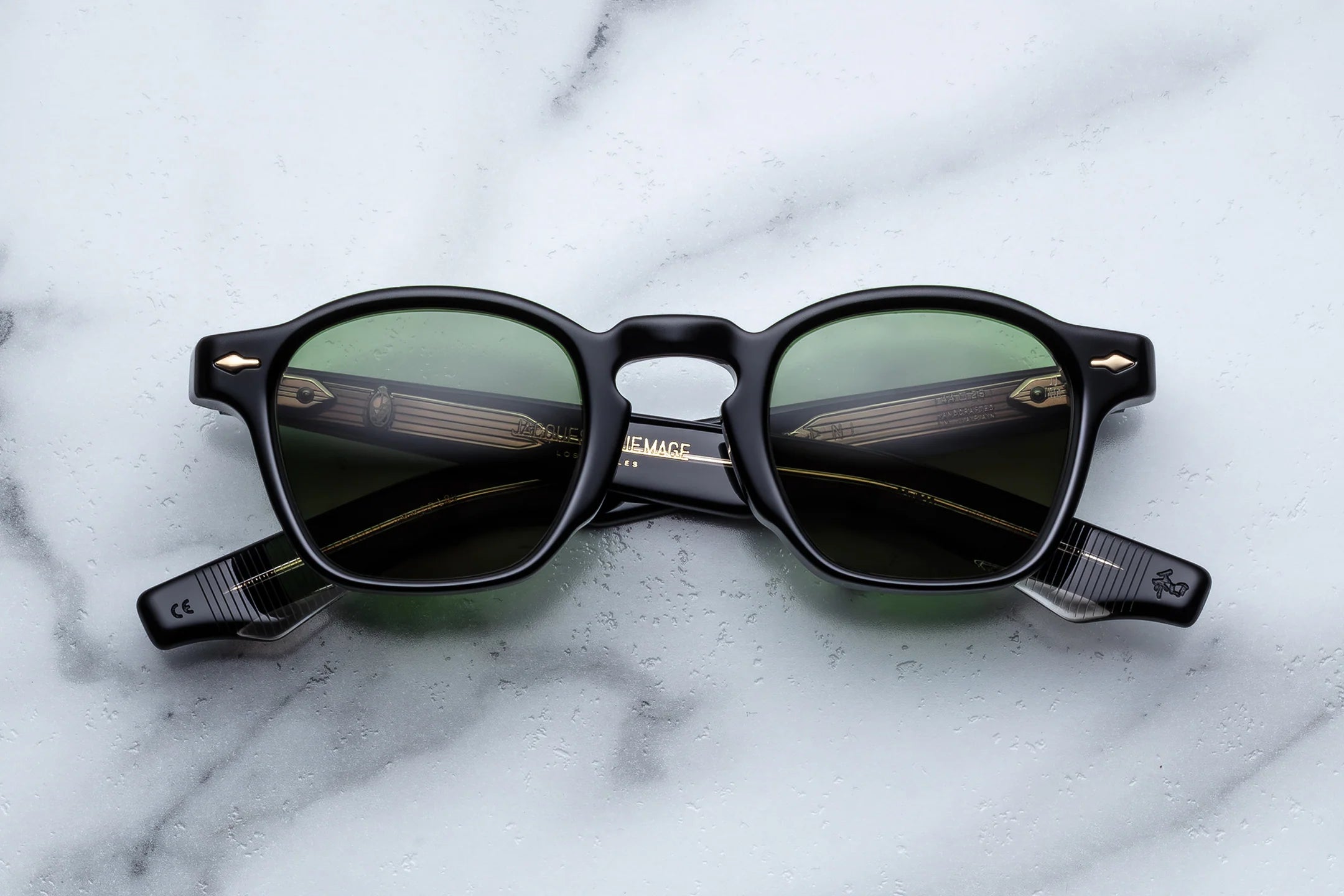 Black acetate sunglasses with green-tinted lenses, gold arrowhead rivets, and exposed wire-core temple detailing, shown front-facing on a light marble surface.