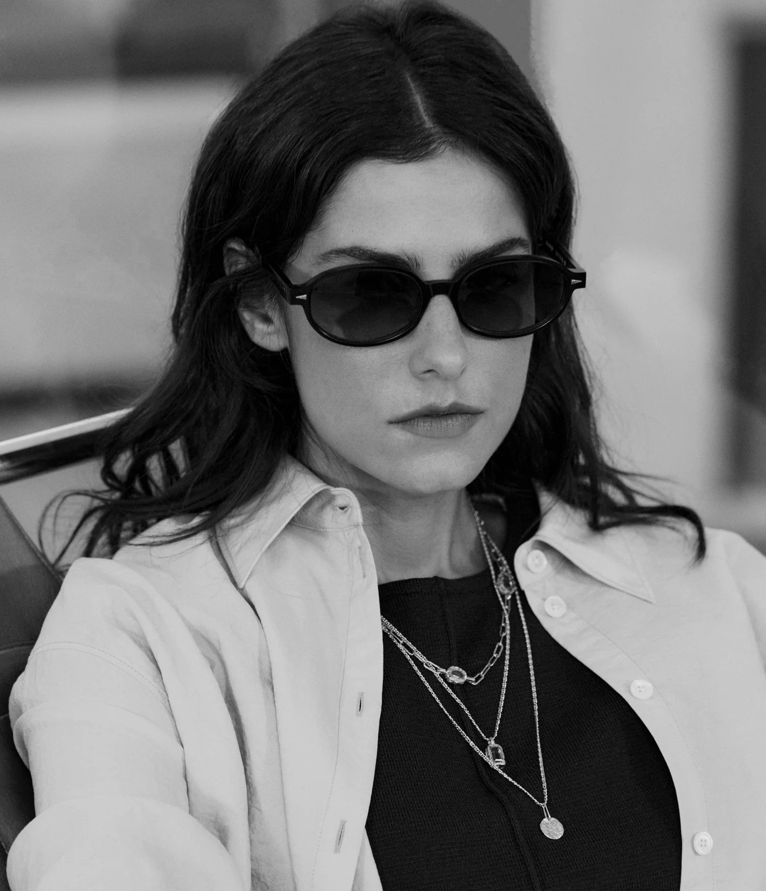 A woman with dark hair wears AHLEM Rohan sunglasses—handcrafted acetate frames by AHLEM, made in France—alongside layered necklaces, a black top, and a light-colored button-up shirt. She looks serious while seated in this black-and-white photo.
