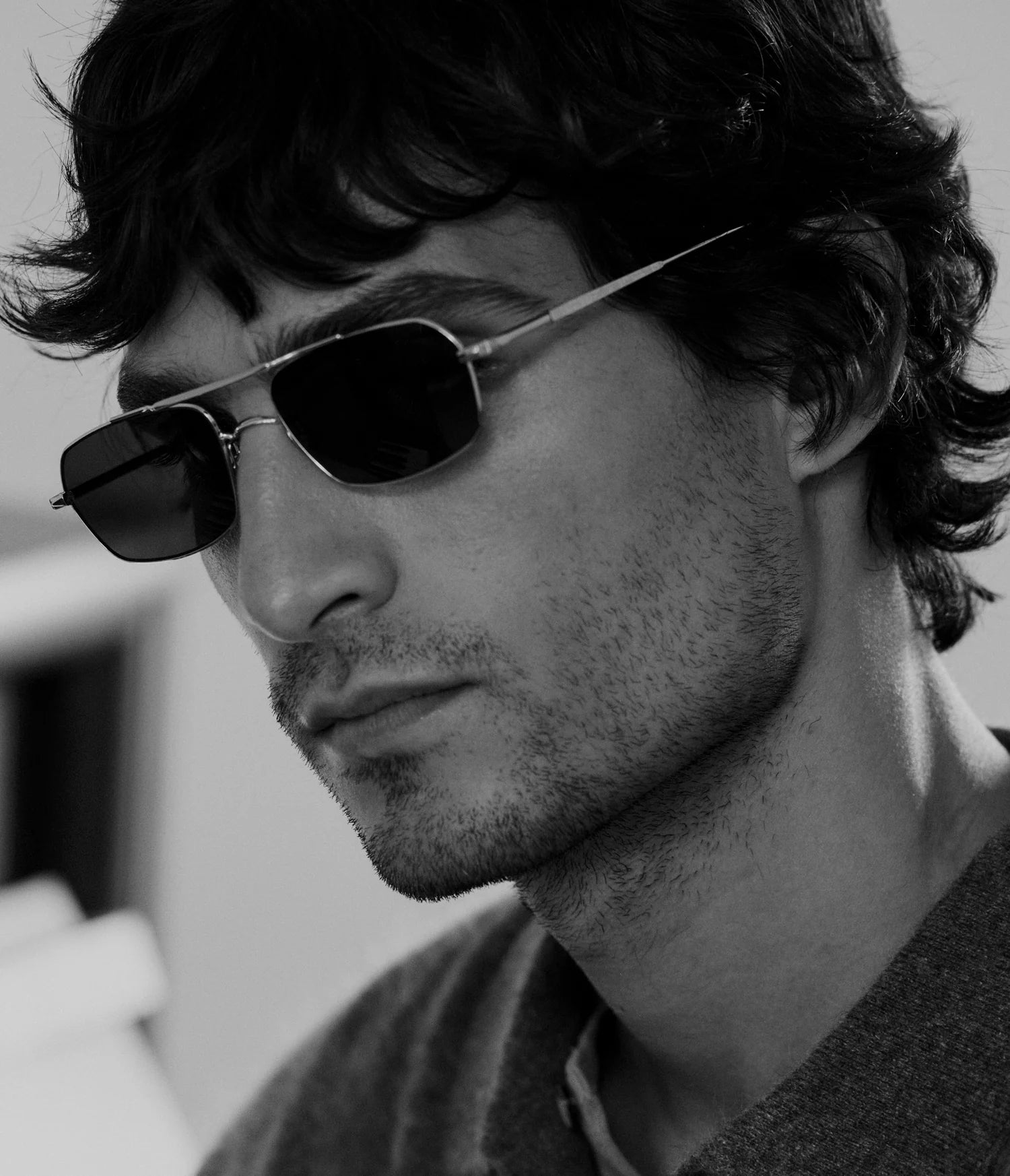A person with wavy hair and light stubble wears AHLEM Trevise rectangular aviator sunglasses—handcrafted in France by AHLEM—and a sweater, looking thoughtful. The image is in black and white.