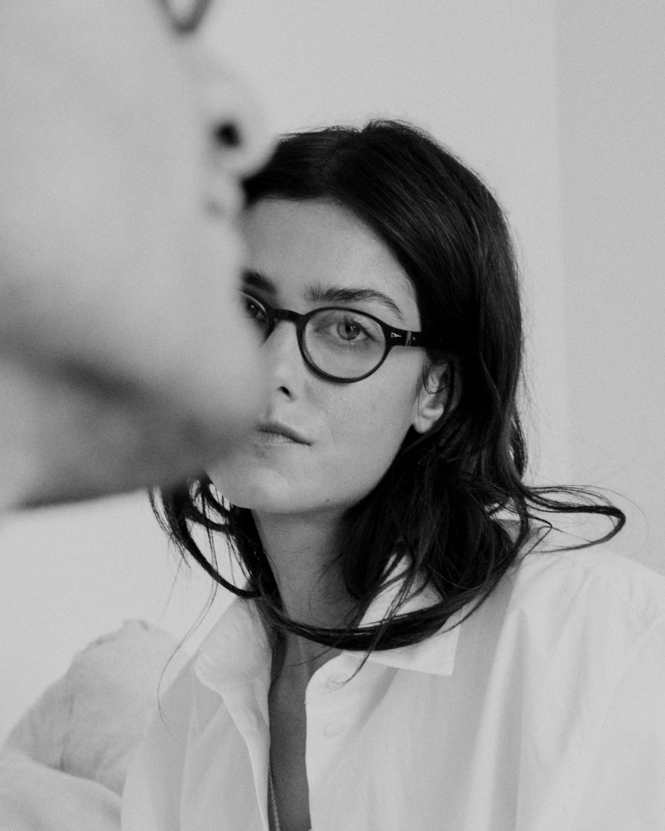 A woman with dark hair and glasses looks at the camera, wearing a white shirt. She wears the AHLEM St Marcel by AHLEM, crafted from premium Italian acetate with 22k gold hardware—its exceptional craftsmanship clear in every detail. Part of another person’s face appears out of focus in the foreground. The image is in black and white.