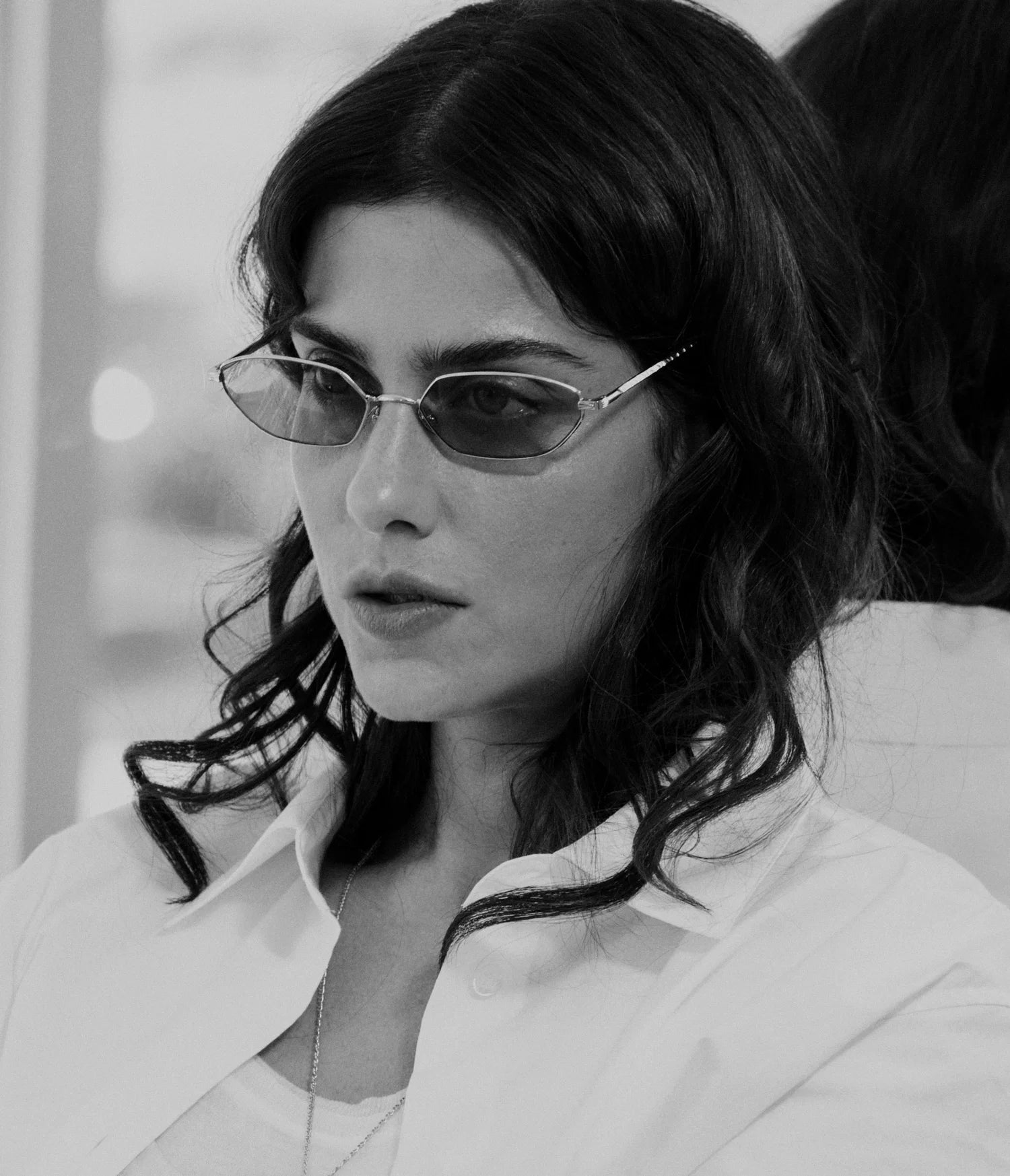 Black and white photo of a woman with wavy hair wearing AHLEM Iris geometric sunglasses by AHLEM, handcrafted in France. She has a serious expression and is facing slightly to the left, dressed in a white shirt with a reflective surface behind her.
