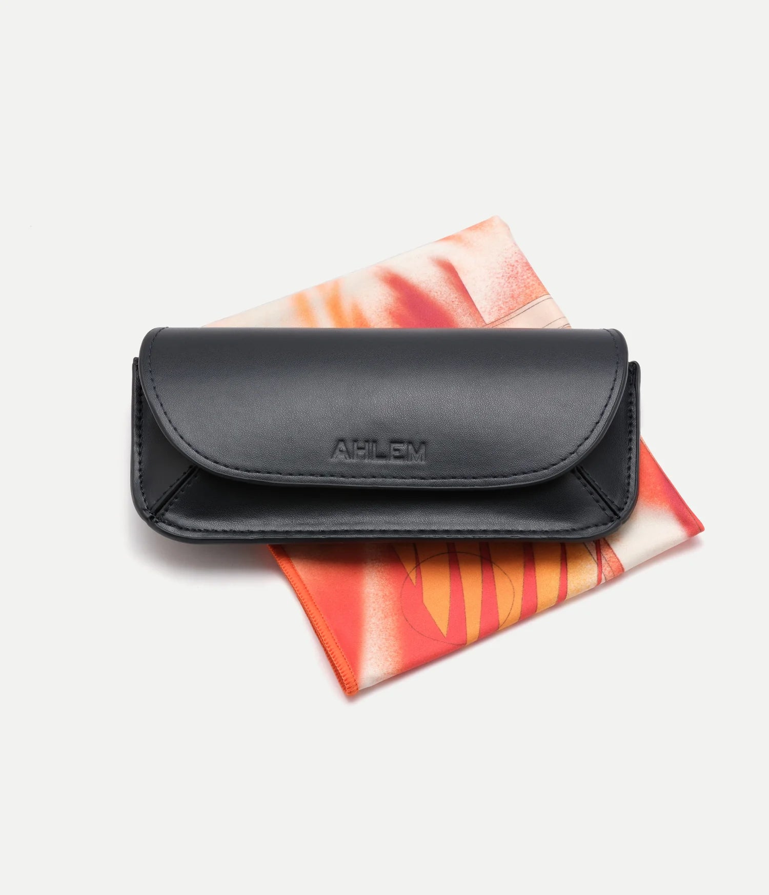 A black leather AHLEM Iris eyeglasses case, embossed with the AHLEM logo on the front, rests atop a vibrant orange and red patterned cloth—an elegant accessory that reflects AHLEM’s tradition of handcrafted French eyewear.