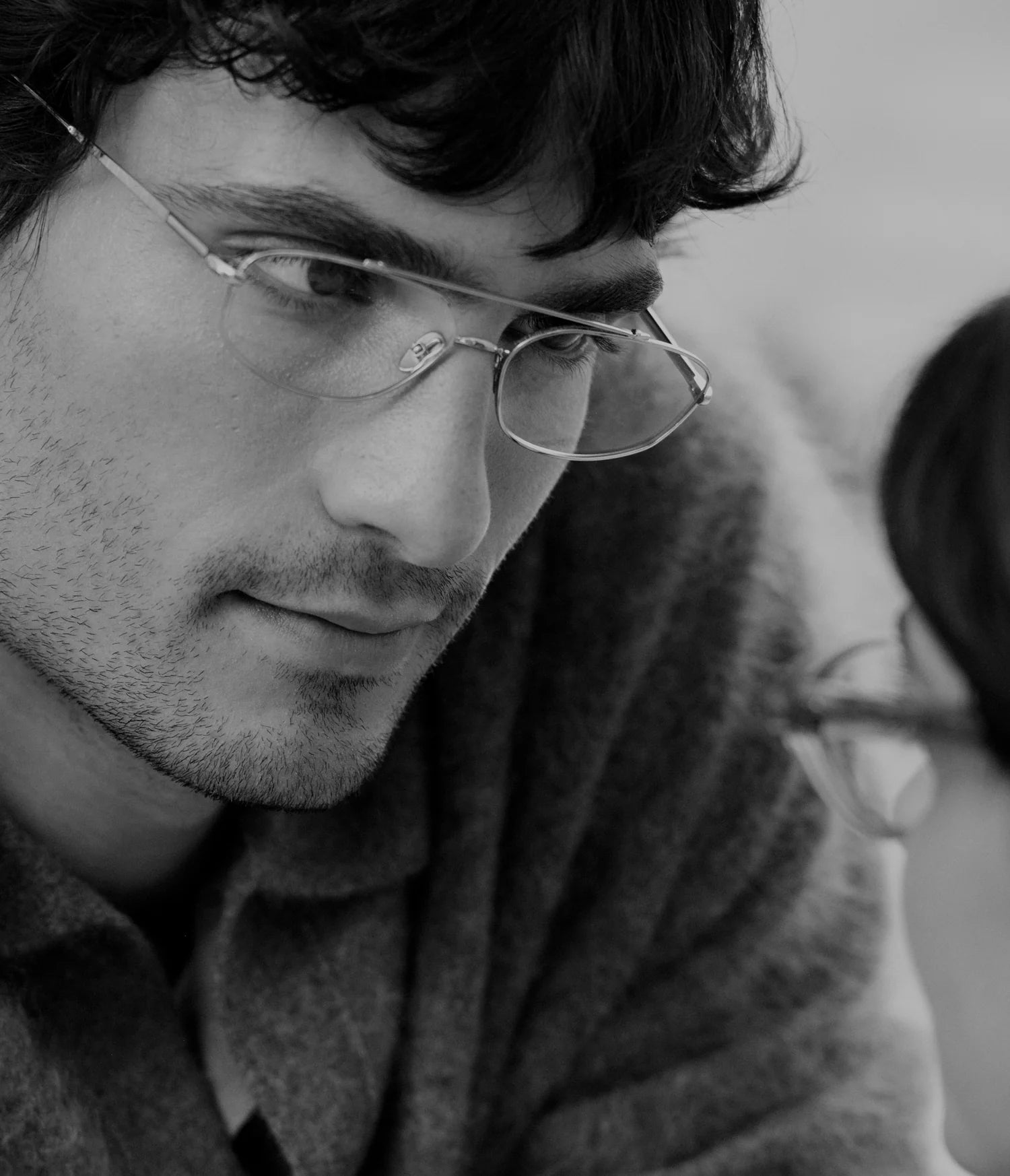 A close-up black and white photograph of a man wearing thin metal rimless eyeglasses and a textured knit sweater, looking downward with a soft, contemplative expression.