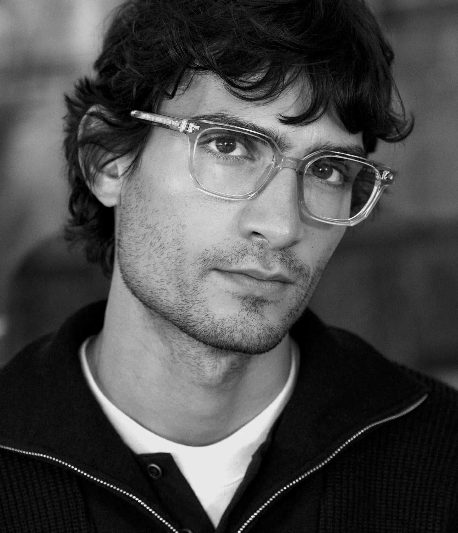 Black and white portrait of a young man with wavy dark hair, light stubble, and clear AHLEM Houdon eyeglasses by AHLEM, wearing a dark zip-up sweater over a white shirt, looking slightly to the side with a neutral expression.