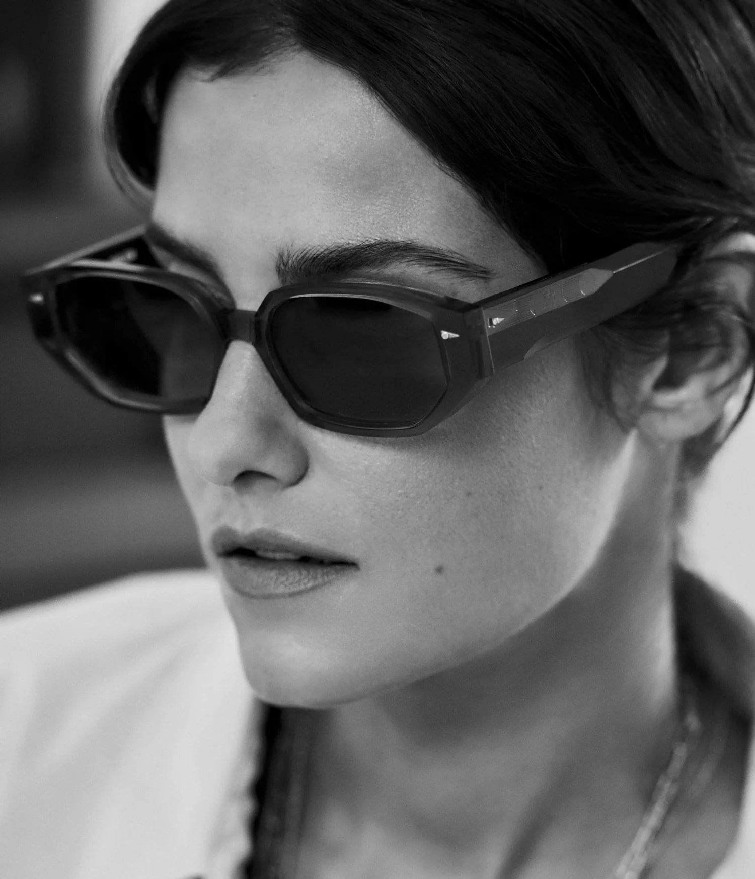 Black and white close-up of a person wearing AHLEM Poitou rectangular sunglasses by AHLEM, featuring dark lenses crafted from premium Italian acetate. They are looking slightly to the side with dark, center-parted hair and a calm expression.