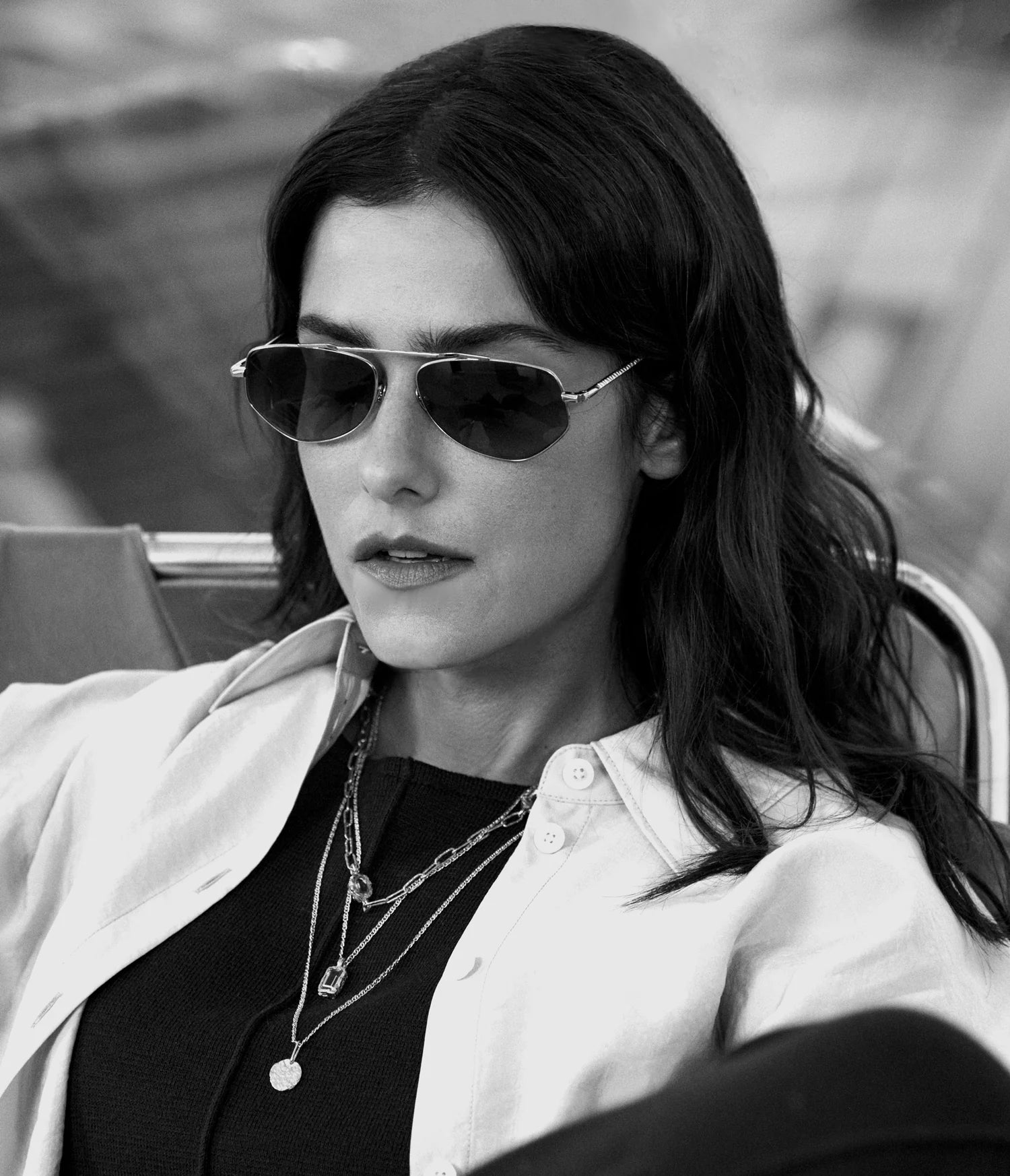 Wearing AHLEM Julien aviator sunglasses by AHLEM, a woman with wavy dark hair sits in a black top and light open shirt, layered in 22k gold necklaces. She looks down with a neutral expression in this black and white photograph.
