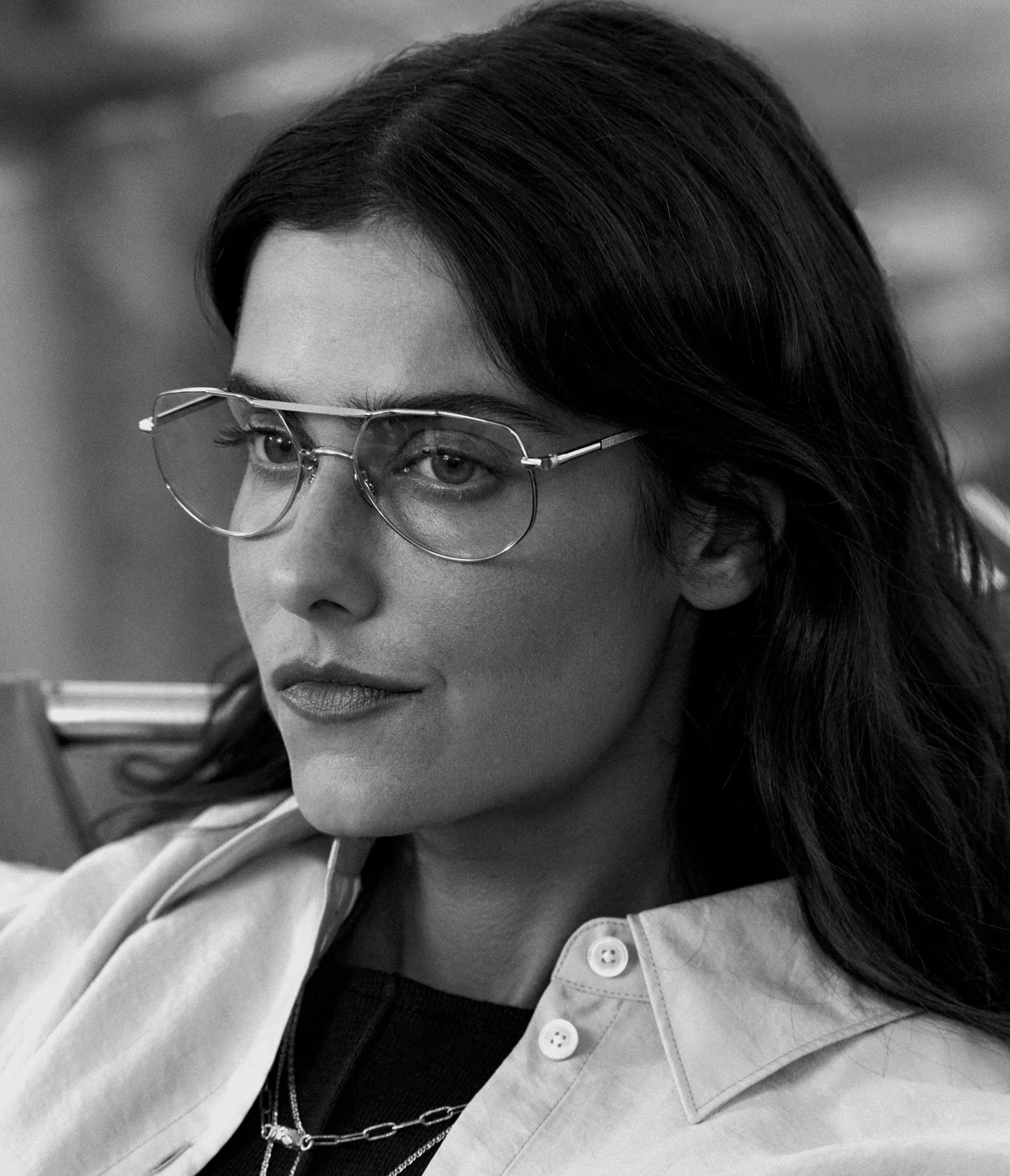 A woman with long hair wears AHLEM Cadet aviator-style glasses by AHLEM, handcrafted in France, along with layered necklaces and a light-colored shirt. She gazes thoughtfully to the side in a black-and-white photograph.