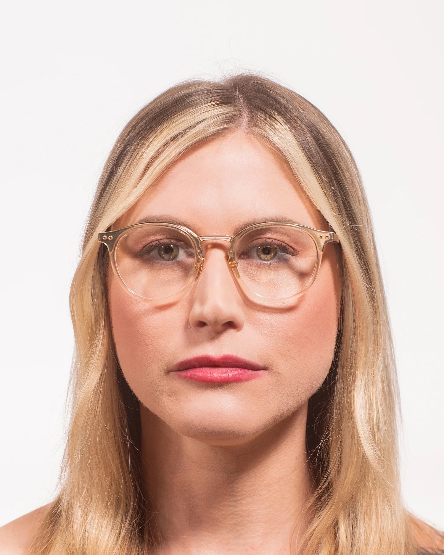 A close-up portrait of a woman with blonde hair wearing round, semi-transparent gold-framed eyeglasses against a plain white background.