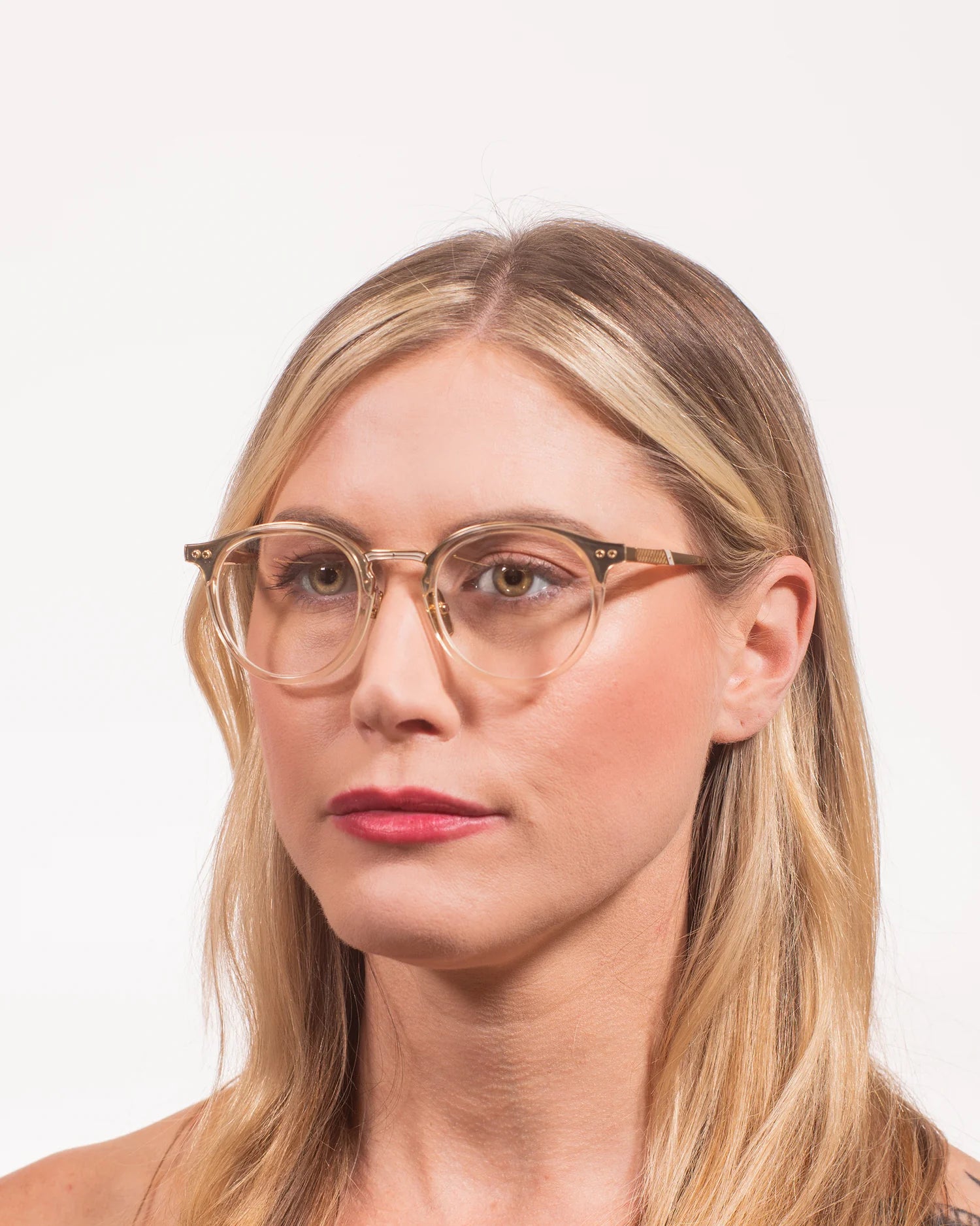 A close-up portrait of a woman with blonde hair wearing round, clear-framed eyeglasses with a thin gold metal rim and small gold geometric accents on the temples, set against a plain white background.