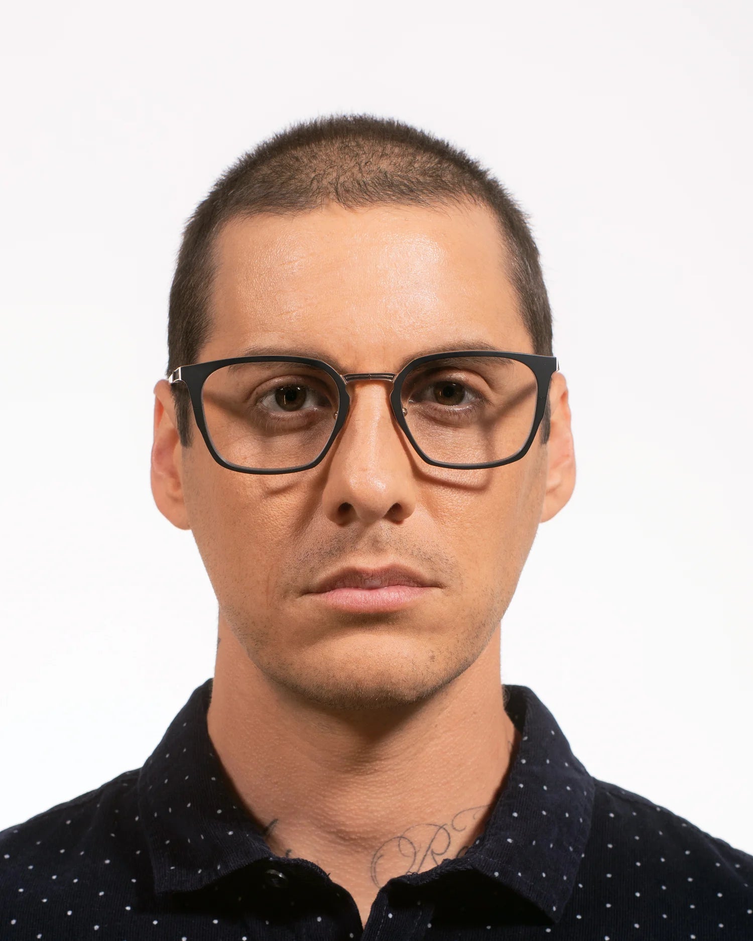 A man with a buzz cut wearing black square-frame eyeglasses and a dark navy polka-dot collared shirt, photographed against a plain white background.
