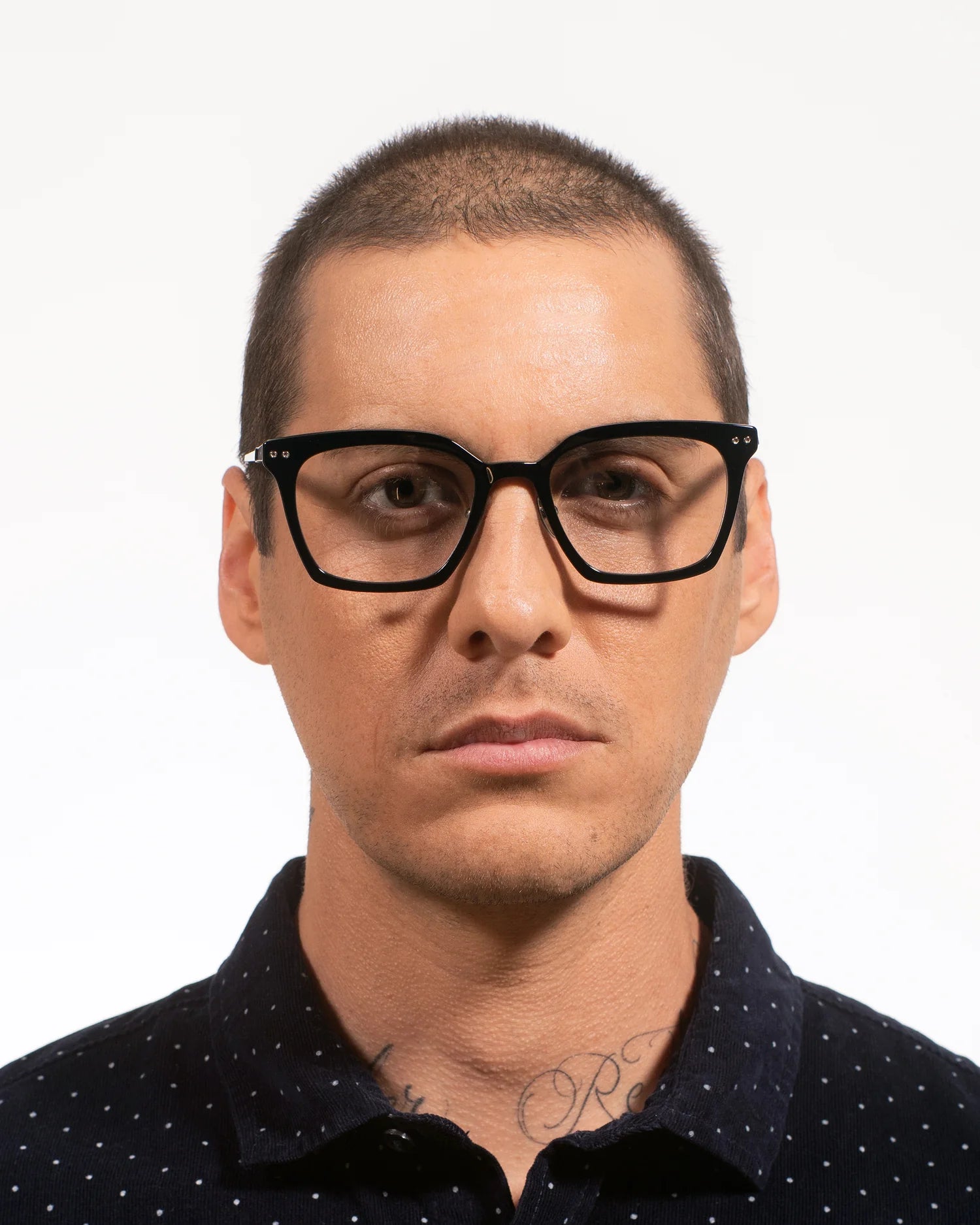 A man with a buzz cut wearing thick black rectangular eyeglasses and a dark navy polka-dot polo shirt, photographed against a plain white background.