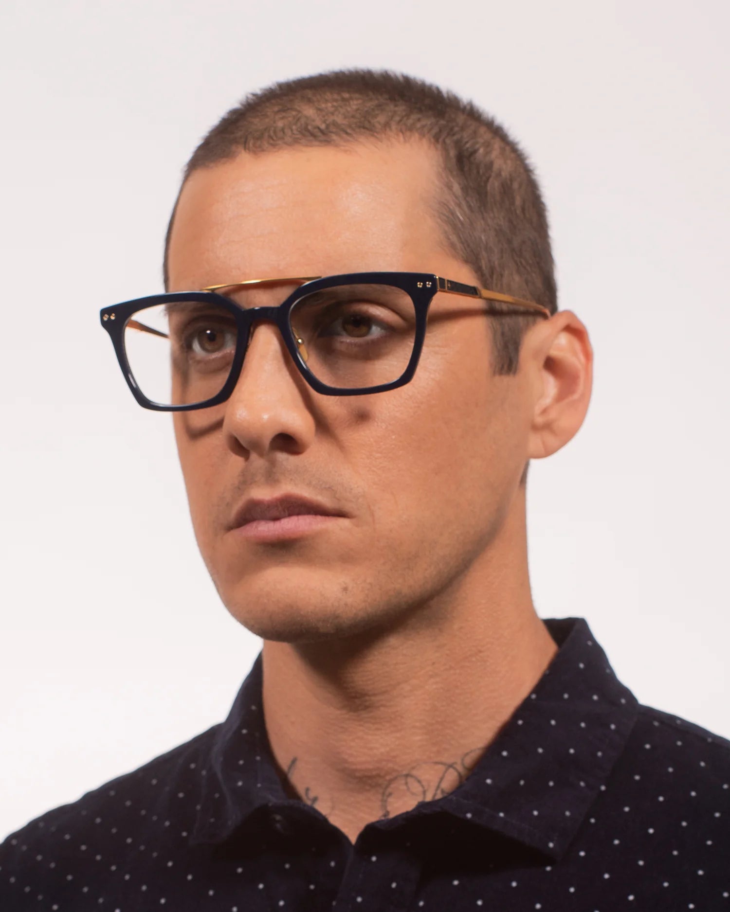 A close-up portrait of a man with a buzz cut wearing thick black square eyeglasses with gold metal temples and clear lenses, against a plain light background.