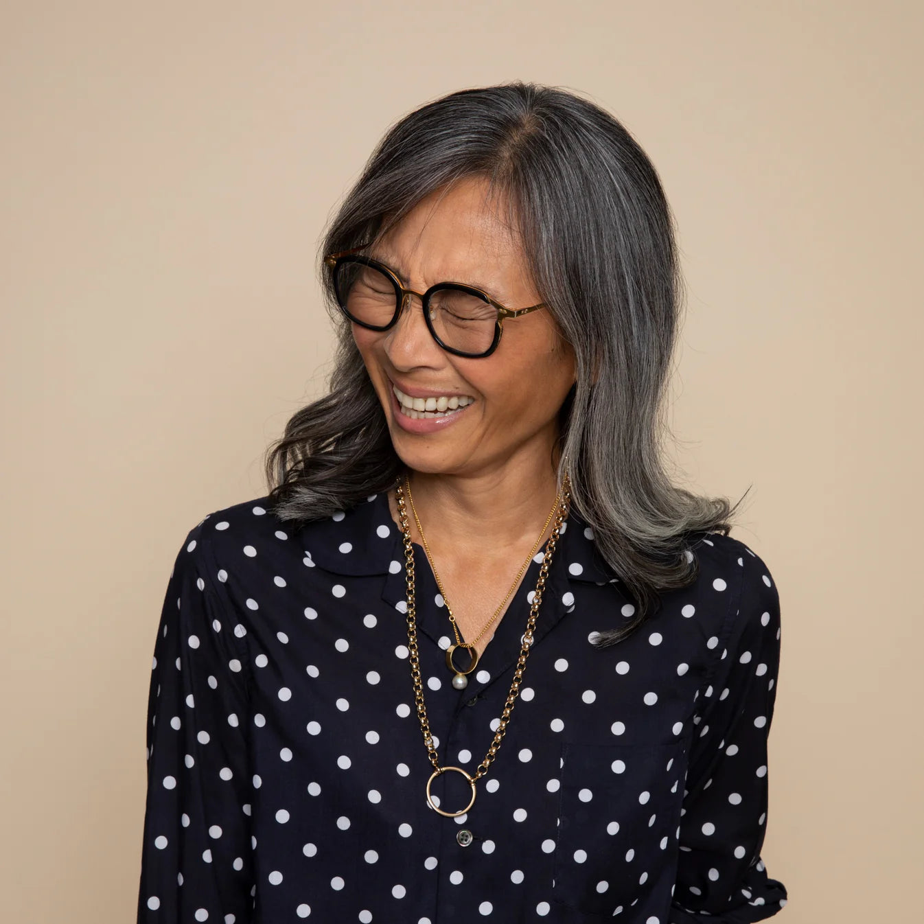 Older woman with long gray hair in a navy polka dot shirt and gold glasses chain smiles with eyes closed against a plain beige background, accessorizing her glasses with the elegant La Loop Camilla Gold Long by La Loop.